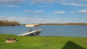 A lakeside deck with a covered seating area, a fire pit, and a grassy lawn.