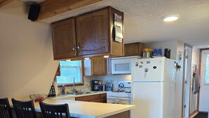A kitchen with wooden cabinets, a white refrigerator, a microwave, and a stove. There are three black chairs around a wooden island.