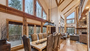 A spacious dining area with a wooden table and chairs, large windows, and a stone fireplace. - Escape Lodge - Hot Tub, Large Deck, Sauna, Gym. (Sundance)