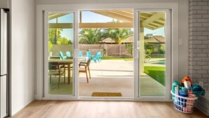 Una habitación con puerta de cristal corredera que da a un patio al aire libre con mesa y sillas, un cesto de toallas y un muro de ladrillo.