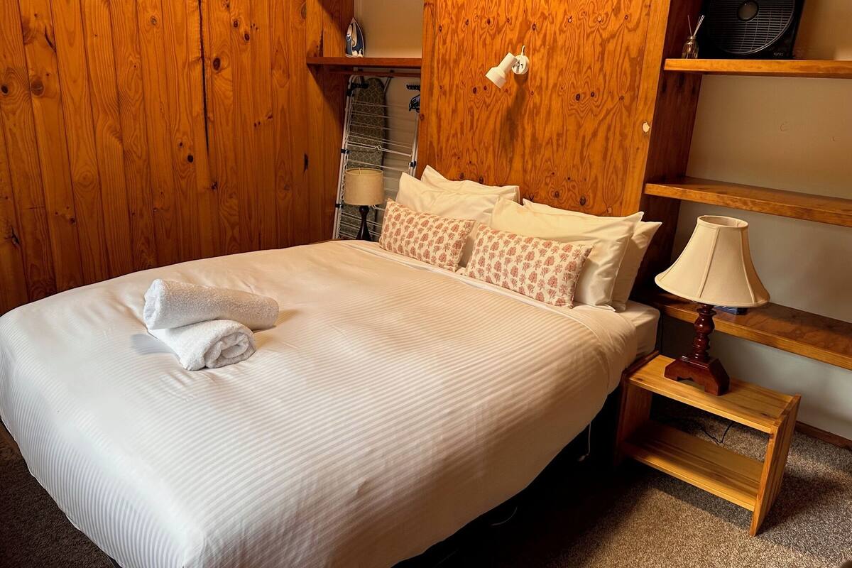 A neatly made bed with a folded towel, wooden shelving with a fan and a lamp, and a small table with a lamp.
