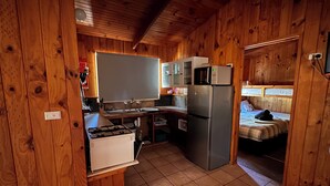 A compact kitchen with a stove, sink, and refrigerator, wooden walls, and a small sleeping area with a bed. - Woodbine Park Eco Cabins (Bournda)