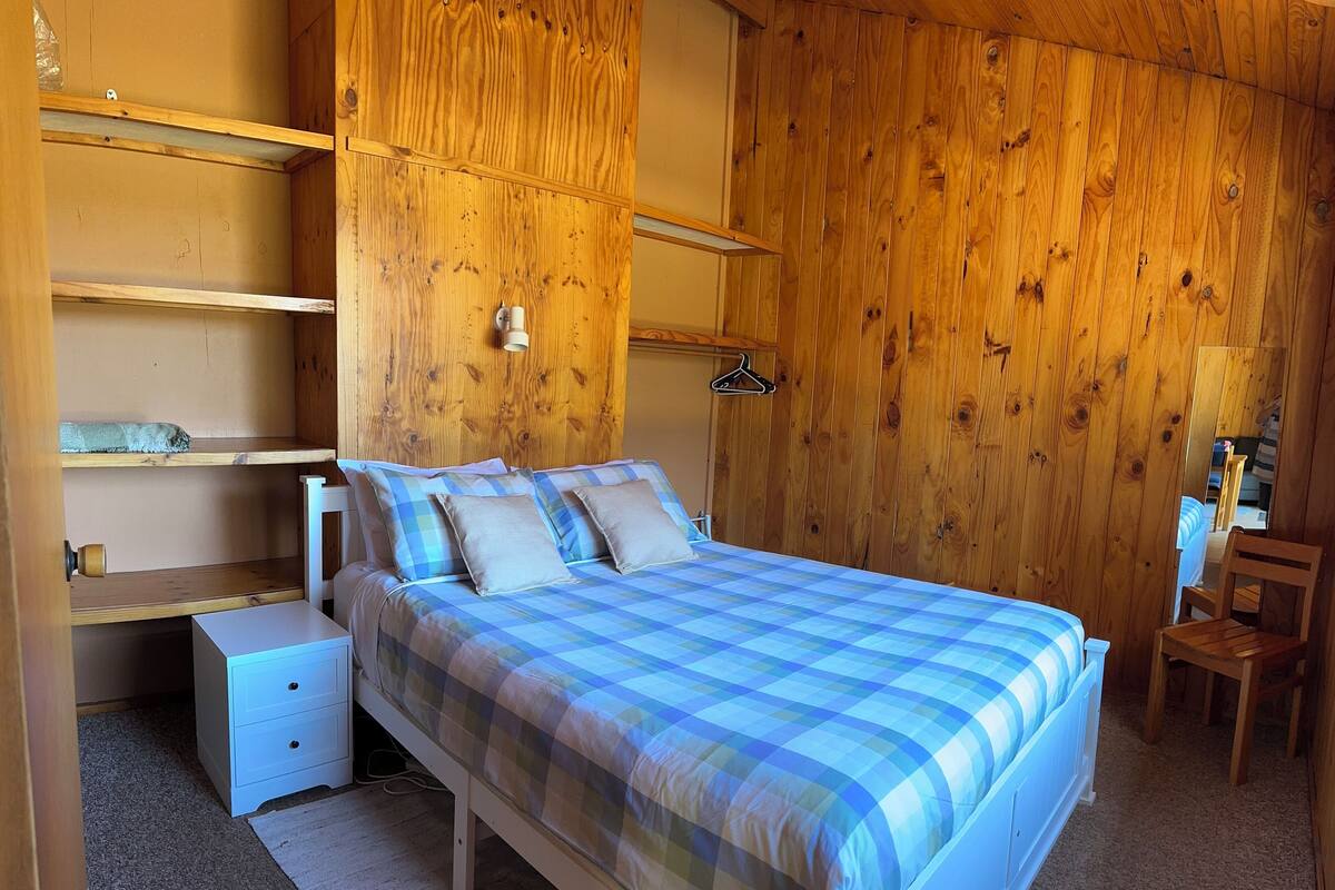 A bedroom with a wooden wall, a bed with a checkered bedspread, a nightstand, a dresser, and a hanging clothes rack.