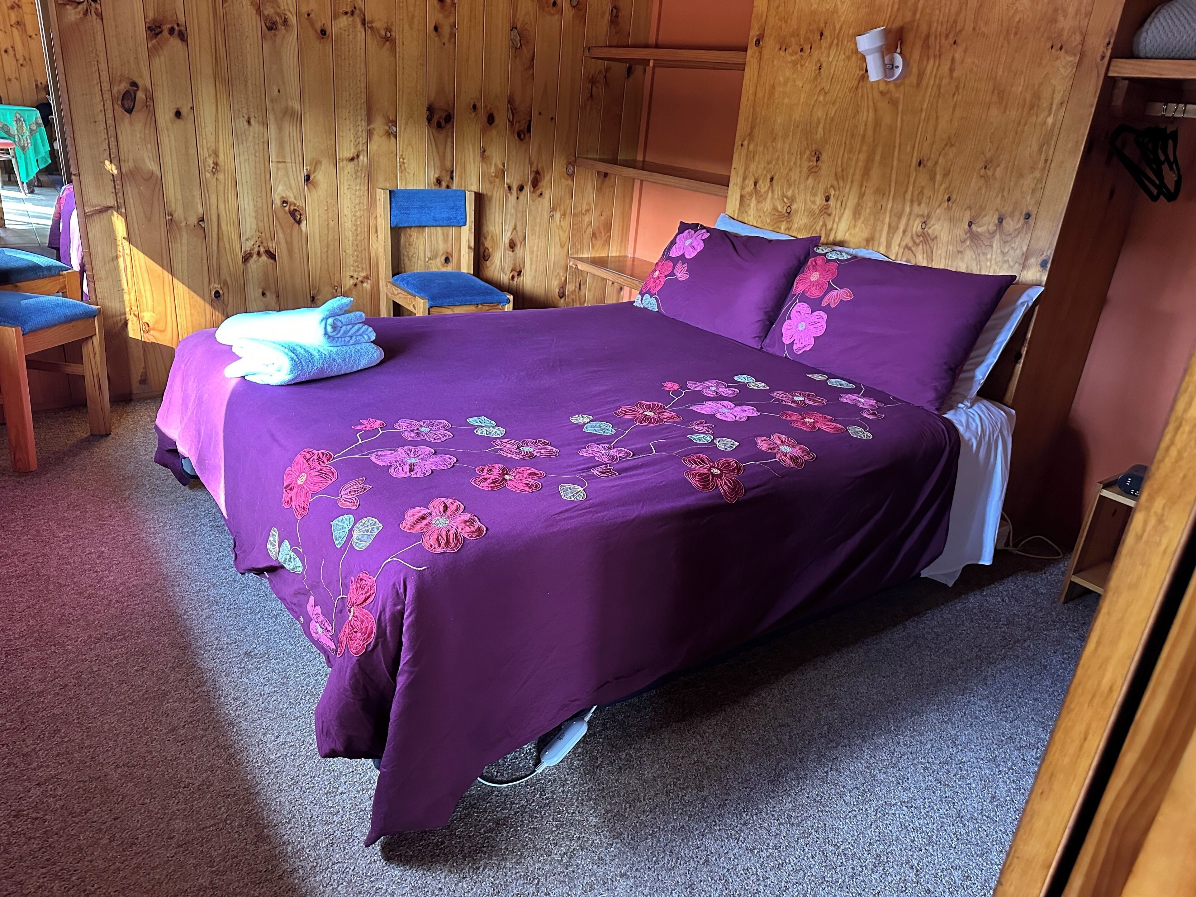 A room with a wooden interior, a bed with purple bedding, a chair, and a small table.