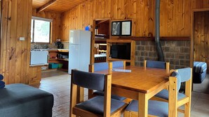 A wooden cabin interior with a dining table, chairs, a kitchen area, a fireplace, and a television. - Woodbine Park Eco Cabins (Bournda)