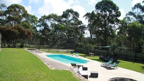 A rectangular swimming pool with a stone border, surrounded by a grassy lawn and trees. There are lounge chairs and a small hot tub nearby. - Woodbine Park Eco Cabins (Bournda)