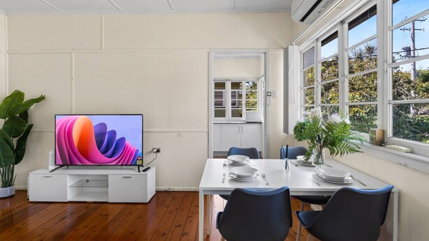 Un comedor moderno con un televisor de pantalla plana grande, piso de madera, paredes blancas y un ventanal con vistas a zonas verdes.