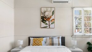 Une chambre à coucher comprenant un lit, deux tables de chevet, une fenêtre avec stores, un climatiseur fixé au mur et, au-dessus du lit, une reproduction encadrée d’une illustration botanique.