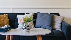 Un canapé noir avec trois coussins, un vase blanc avec des fleurs et une boîte de mouchoirs sur une table blanche.
