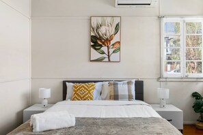 A bedroom with a bed, two bedside tables, a window with blinds, a wall-mounted air conditioner, and a framed botanical print. - Cozy 3BR Home in Yeronga | Garden & Outdoor Living (Yeronga)