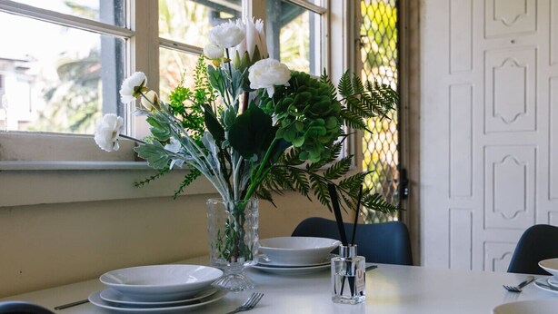 Une table à manger dressée avec des assiettes, des couverts et un centre de table composé de fleurs blanches et de feuillage vert.