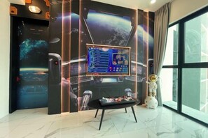 A room with a large window, a wall-mounted screen displaying a game, a small table with a remote control, and a decorative astronaut figure. - Alfa Bangsar by The Homez (Kuala Lumpur)