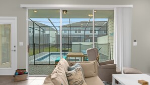Una sala de estar moderna con un sofá color beige, una mesa de centro blanca y un ventanal que da a un jardín con piscina y mesa de patio.