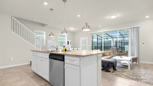 Una cocina moderna con armarios blancos, lavavajillas integrado y encimera de granito. Hay un comedor con mesa y sillas blancas, y un ventanal con vistas al exterior.