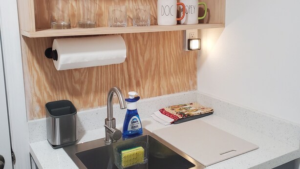 Una cucina con un lavello, una mensola in legno sopra e un piano di lavoro con un portaoggetti per piatti e una bottiglia di detersivo.
