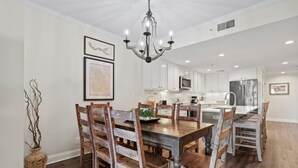 Un comedor amplio con una mesa y sillas de madera, un candelabro y armarios blancos en la cocina contigua.