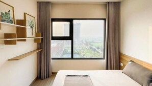 A hotel room with a bed, a window with curtains, a shelf with framed pictures, and a view of buildings. - It Dust Homestay 6 - the Forest Apartment (Hanoi)