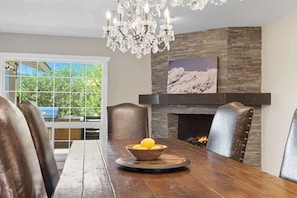 A dining area with a wooden table, leather chairs, a chandelier, a fireplace, and a window with a view of greenery.