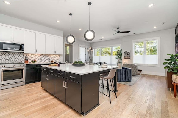 A modern kitchen with a large island, stainless steel appliances, pendant lighting, and a view of the outdoors.