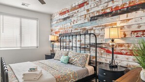 Une chambre à coucher avec un lit en métal, une couvre-lit à motifs, deux tables de chevet avec des lampes et une fenêtre avec des stores.