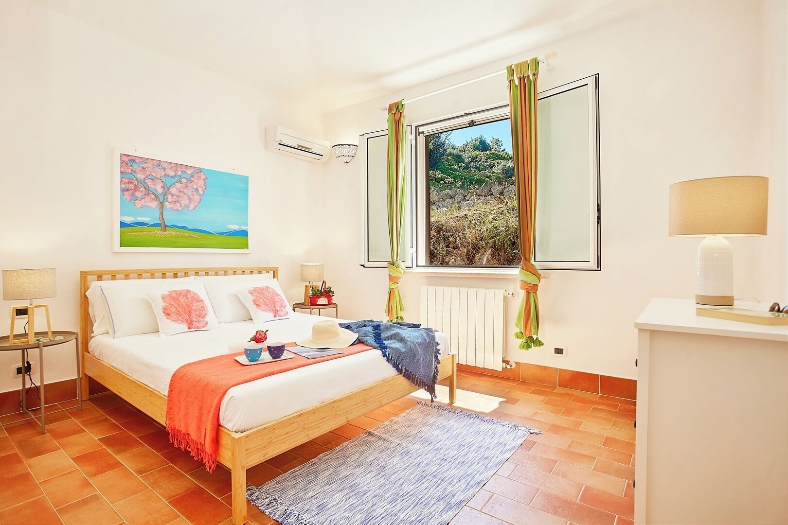 Een slaapkamer met een bed, nachtkastje, raam met gordijnen, radiator en een schilderij aan de muur.