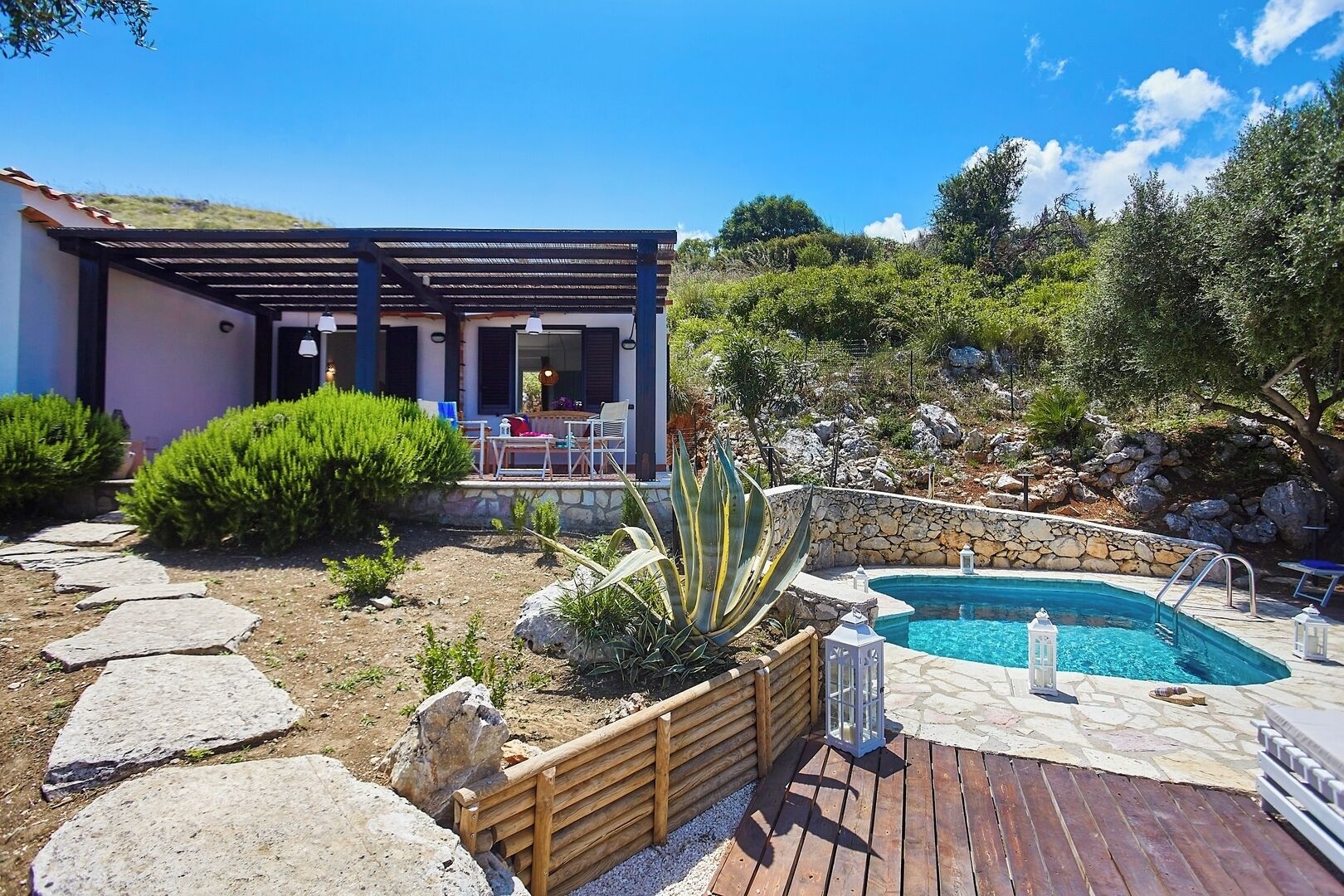 Un área de piscina con cubierta de madera, pared de piedra y pérgola con mobiliario exterior.