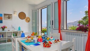 A bright room with a dining table set for breakfast, a bookshelf, and a view of the sea. - Villa Didi – Scopello, Castellamare del Golfo area (Scopello)