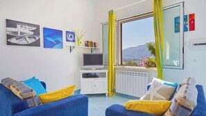 Moderni olohuone, jossa on suuri ikkuna, litteänäyttöinen televisio ja taideteoksia seinillä.