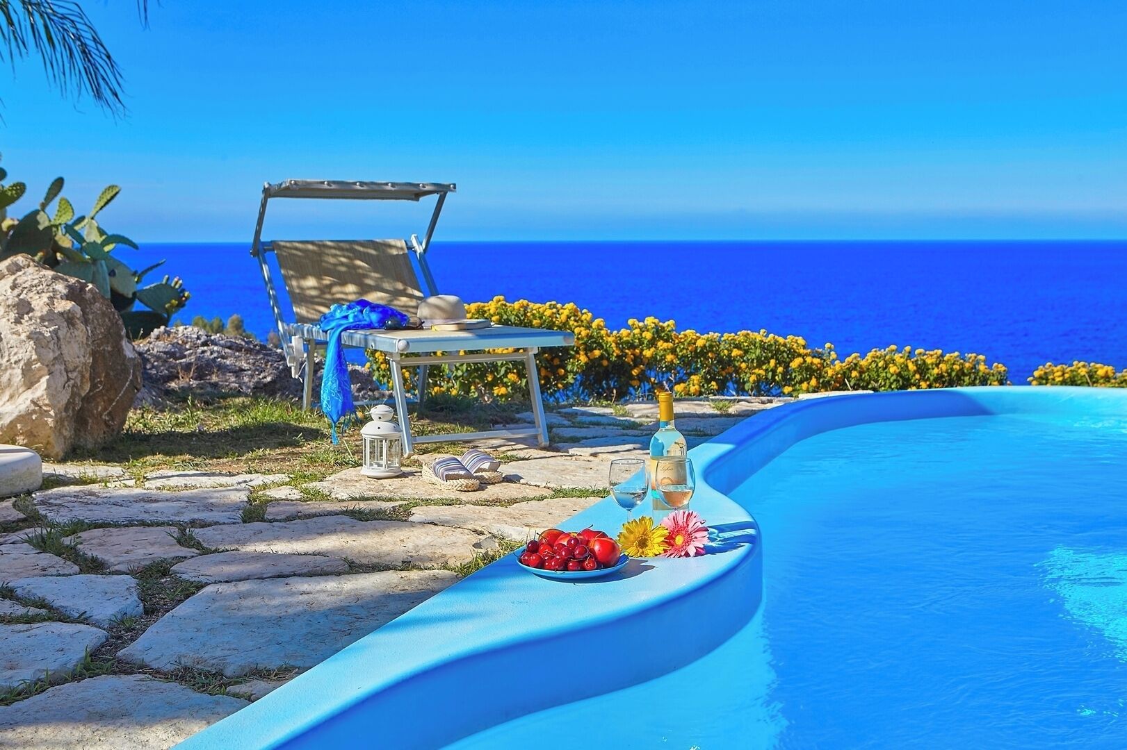 Un espace aménagé au bord de la piscine, avec un fauteuil de relaxation, une table avec des boissons et une vue sur l’océan.