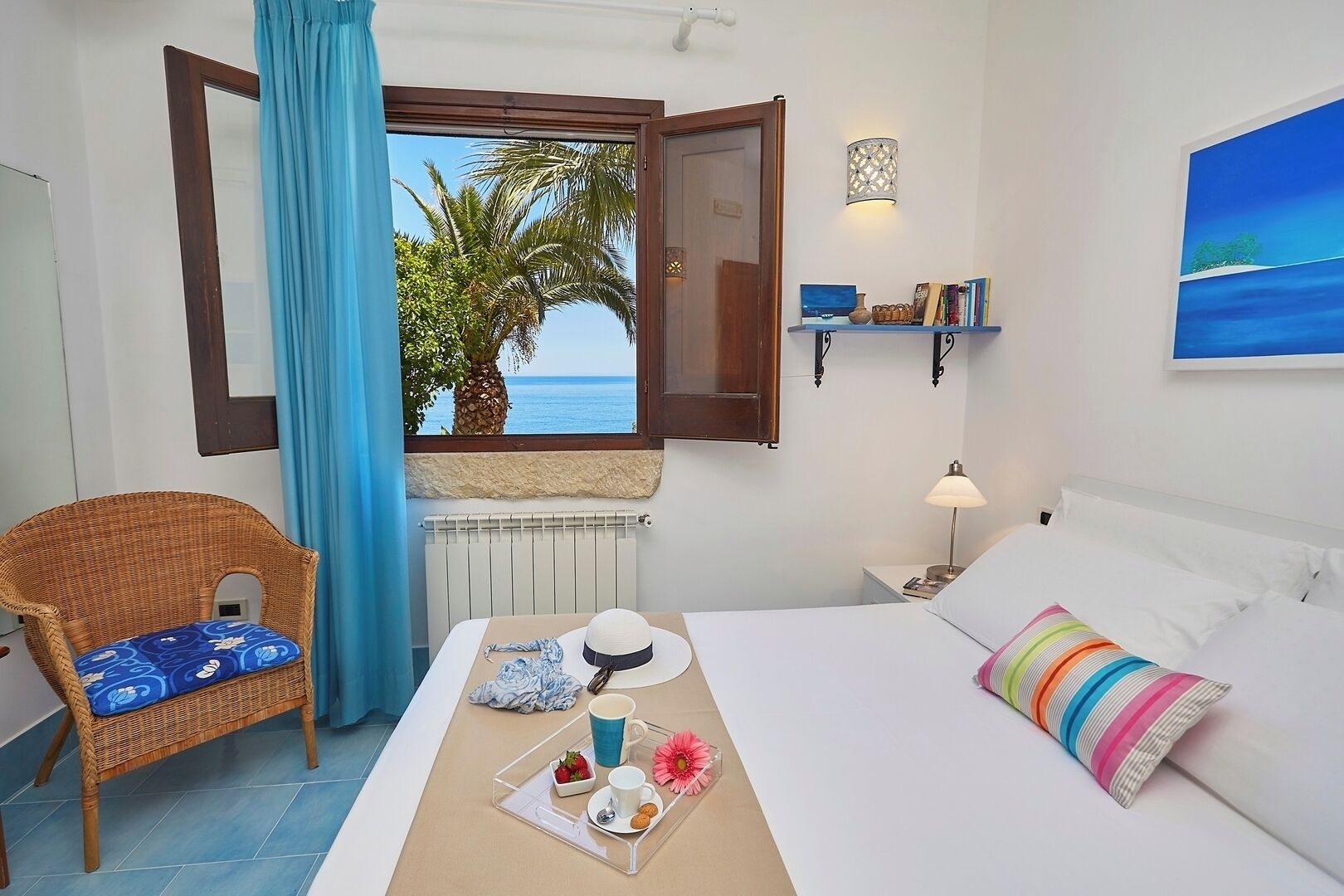 Une chambre d’hôtel avec un lit, une chaise en osier, une table à manger avec des articles pour le petit-déjeuner, une vue sur la mer et une étagère avec des livres et des objets décoratifs.