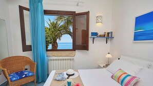 Habitación de hotel con cama, una silla de mimbre, una mesa de comedor con alimentos para el desayuno, vista al mar y una estantería con libros y objetos decorativos.
