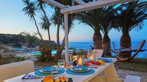 Mobiliario de comedor al aire libre con vista al mar, palmeras y sillones de descanso.