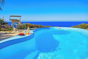 A curved infinity pool with a clear blue ocean view, a sun lounger, and a table with fruits and a bottle. - Villa Sicilia, near the sea (Scopello)