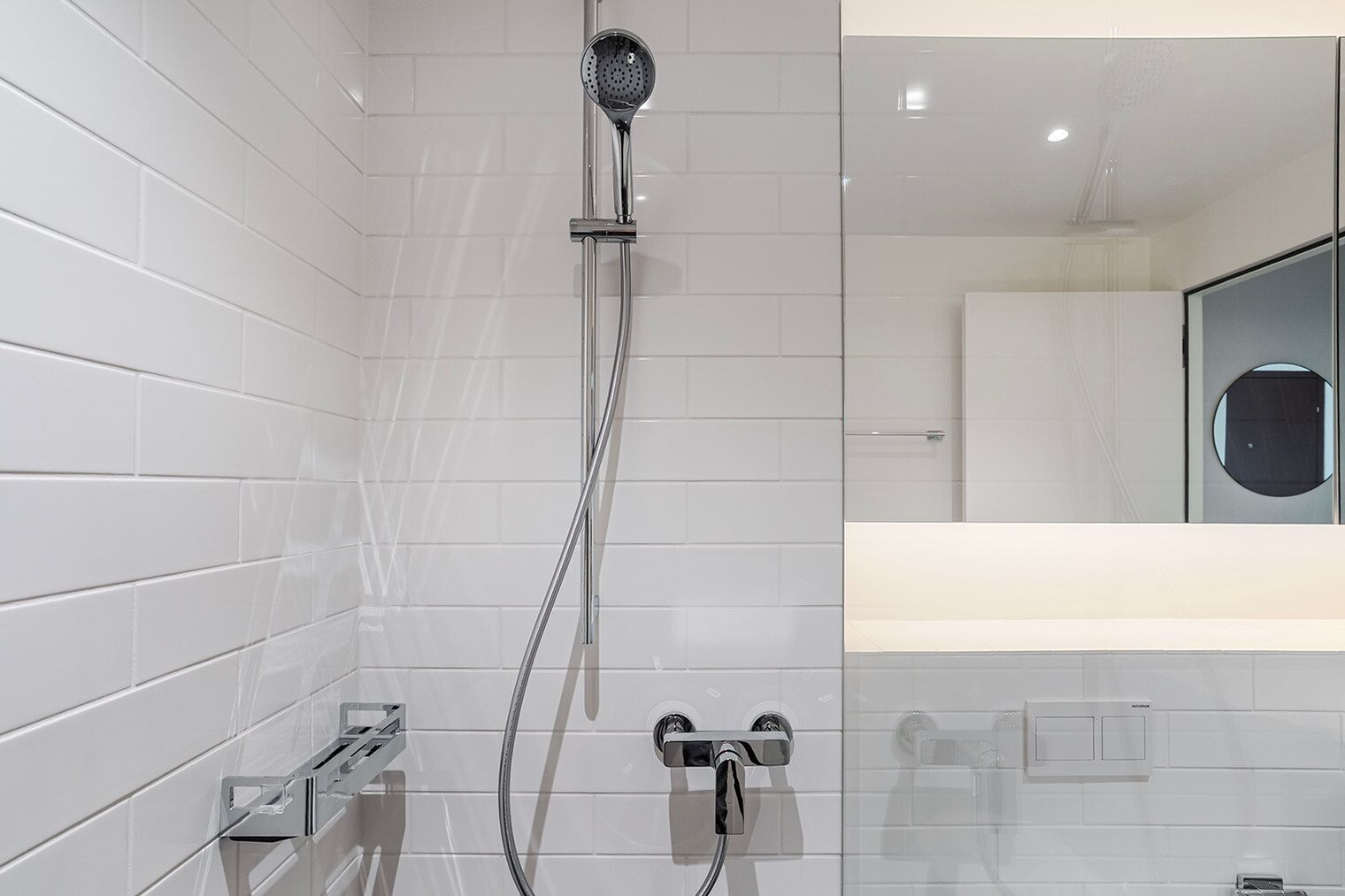 Un baño moderno con paredes de azulejos blancos, ducha con cabezal de cromo y espejo visible.