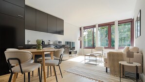 A modern kitchen with dark cabinets, a dining area with a wooden table and chairs, and a living area with a sofa and a lamp. - Blueground | Basel-Vorstaedte, view, nr Kunstmuseum (Basel)
