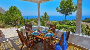 An outdoor dining area with a table set for four, overlooking a scenic view of the sea and hills.