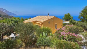 A yellow building with a tiled roof surrounded by lush greenery and cacti, overlooking a clear blue sea.