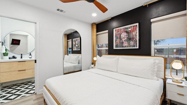Een moderne slaapkamer met een bed, een plafondventilator, een nachtkastje met een lamp, een spiegel en kunst aan de muur.