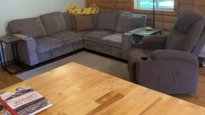 Un salon chaleureux avec un canapé d’angle, un fauteuil inclinable, une table basse en bois, un ventilateur de plafond, une horloge et une fenêtre donnant sur les arbres.