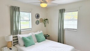Une chambre à coucher comprenant un lit, une table de chevet, une lampe, un ventilateur de plafond et des fenêtres avec des rideaux.