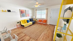 Un salon comprenant un canapé, des fauteuils, une table basse, un tapis, un ventilateur de plafond et une étagère jaune ornée de plantes.