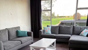 A living room with two grey sofas, a white coffee table, and a large window with a view of a car and greenery outside. - Beach Valley Air B & B (Tuatapere)