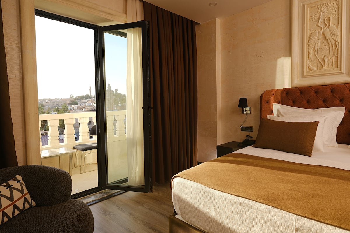 A hotel room with a bed, a nightstand, a lamp, a chair, and a view of a balcony with a cityscape.