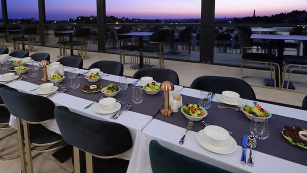 A restaurant with tables set for dining, large windows offering a view of the cityscape at dusk. - Park Dedeman Midyat (Midyat)