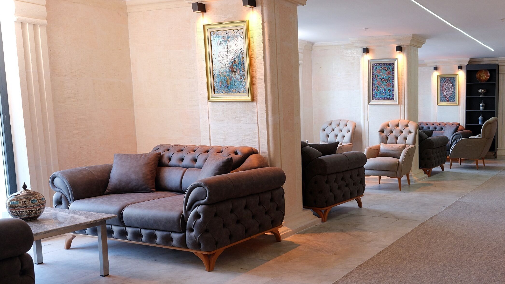 Amplia sala de estar con un sofá de cuero marrón, un sillón a juego y un banco acolchado. La habitación cuenta con techos altos, decoración en las paredes y piso de baldosas.