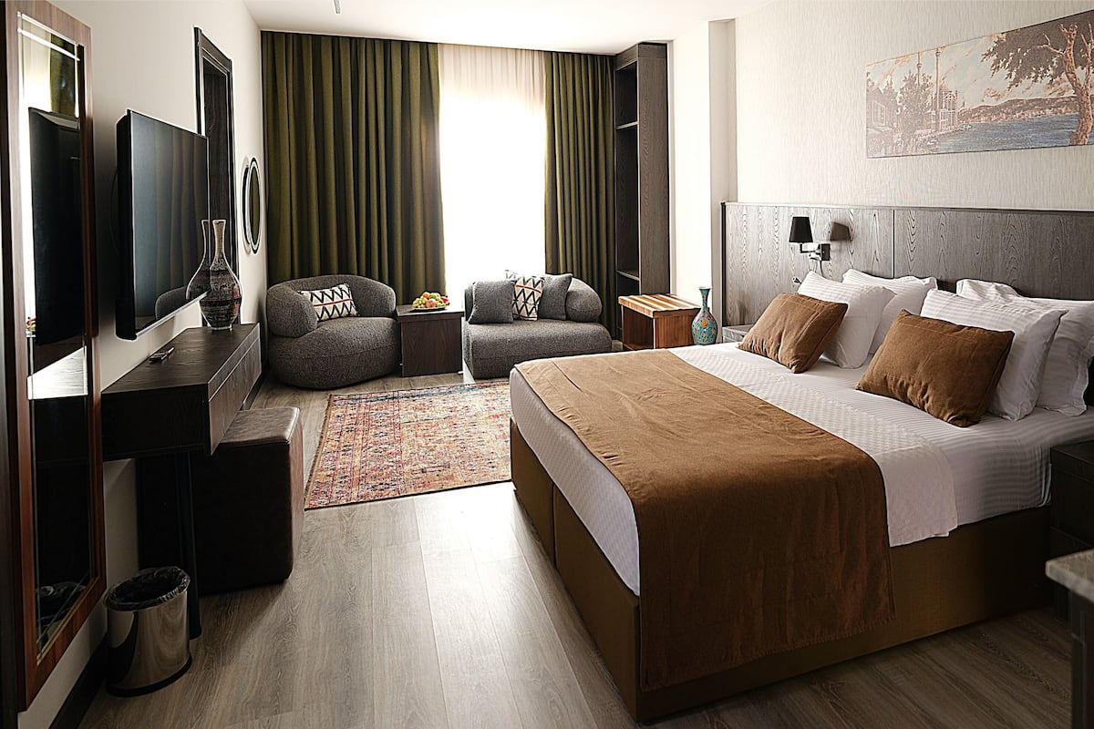 A modern hotel room with a large bed, a seating area with a sofa and armchairs, a TV, and a painting on the wall.