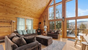 L’intérieur chaleureux d’un chalet, avec un grand plafond en bois, une cheminée et une vue sur l’extérieur grâce à de grandes fenêtres.