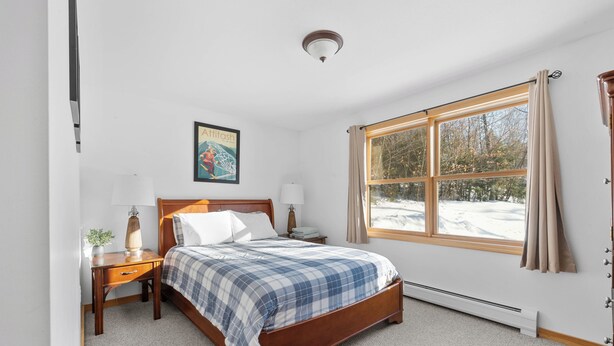 Une chambre à coucher avec un lit en bois, une table de chevet, une lampe, une fenêtre avec des rideaux et un radiateur.