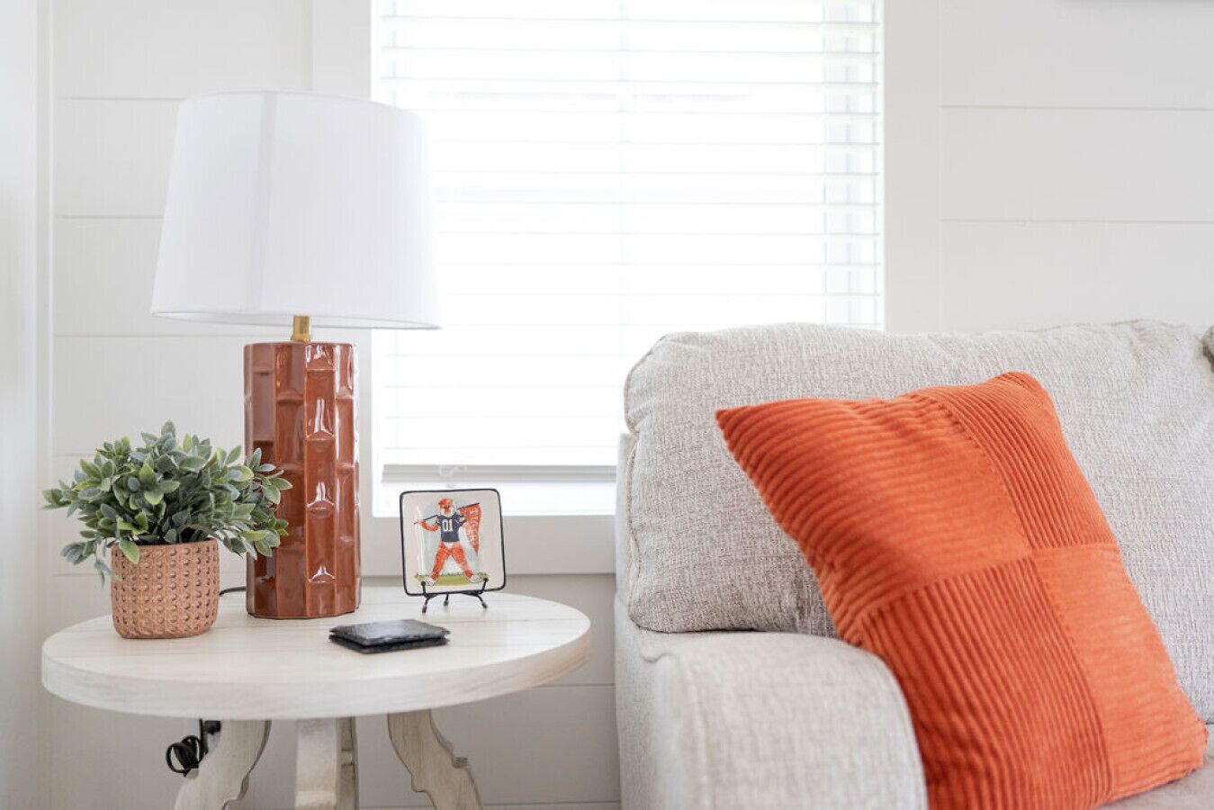 A living room with a white table, a lamp, a potted plant, a framed picture, and a grey sofa with an orange pillow.
