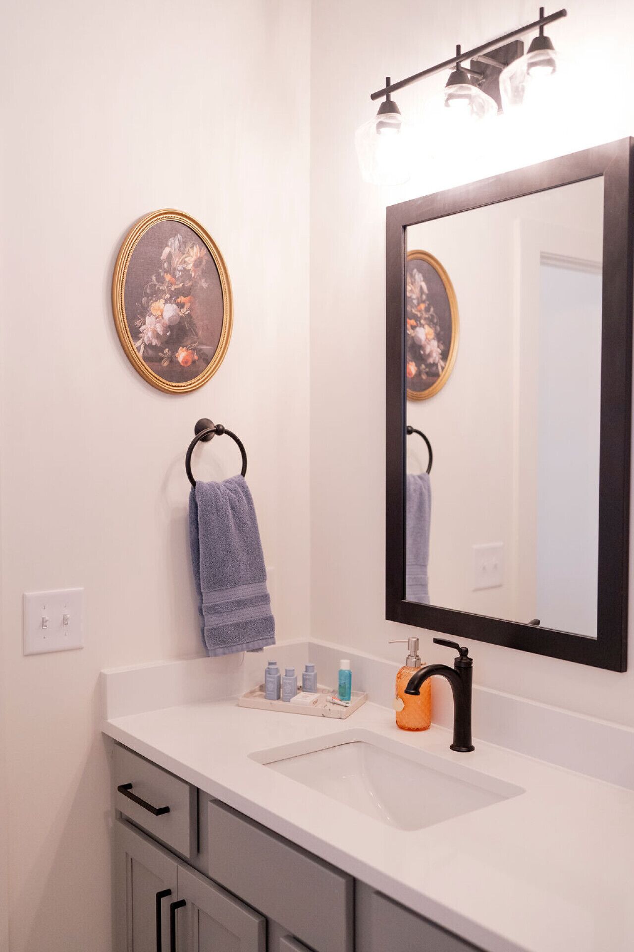 Un bagno con un lavandino bianco, uno specchio, un appendi-asciugamani ad anello e un quadro floreale appeso alla parete.